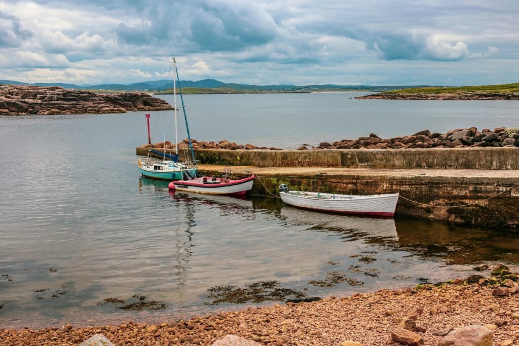 A peaceful harbor scene in Ireland featuring three moored boats against a rocky shoreline under a cloudy sky.