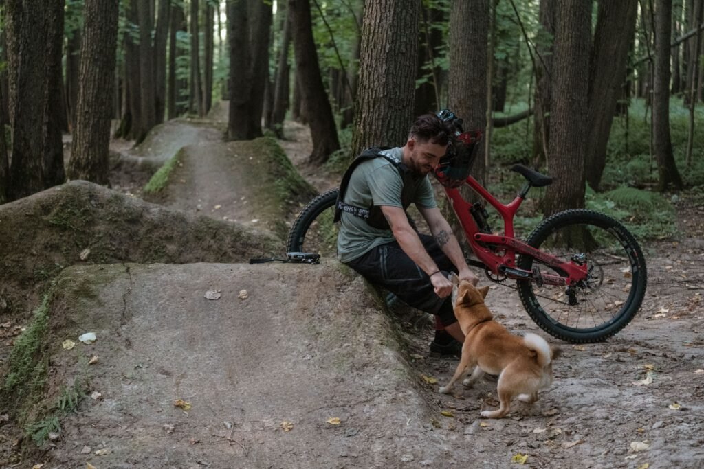 A bearded man sits interacting with a dog next to a bicycle in a peaceful forest setting.